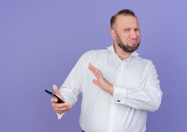 Bärtiger Mann, der weißes Hemd hält, das Smartphone hält Verteidigungsverteidigungsgeste mit Hand, die mit angewidertem Ausdruck steht, der über blaue Wand steht