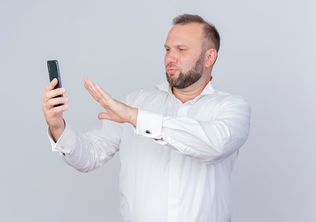 Bärtiger Mann, der weißes Hemd hält, das Smartphone hält Videoanruf, der Bildschirm betrachtet, der Verteidigungsgeste macht, die über weißer Wand steht