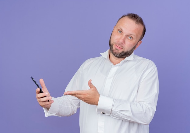 Bärtiger Mann, der weißes Hemd hält, das Smartphone präsentiert, das mit Arm verwirrt über blauer Wand steht