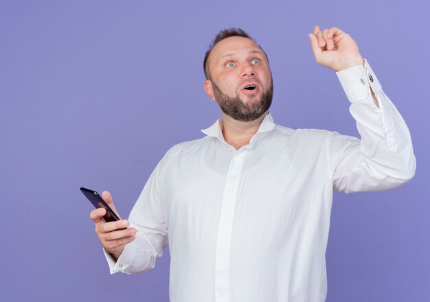 Bärtiger Mann, der weißes Hemd hält, das Smartphone schaut, das überrascht zeigt Zeigefinger, der neue Idee hat, die über blaue Wand steht