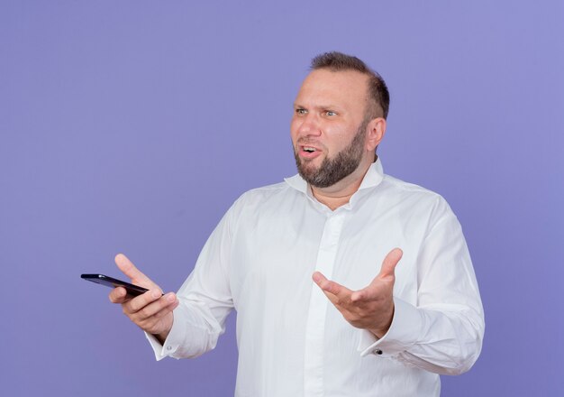 Bärtiger Mann, der weißes Hemd hält, das Smartphone schaut, das verwirrt steht über blauer Wand