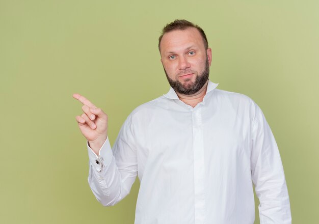 Bärtiger Mann, der weißes Hemd mit ernstem Gesicht trägt, das mit dem Finger zur Seite zeigt, die über heller Wand steht