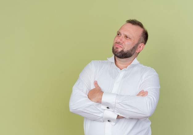 Bärtiger Mann, der weißes Hemd trägt, das mit ernstem Gesicht mit verschränkten Armen auf der Brust über der hellen Wand beiseite schaut