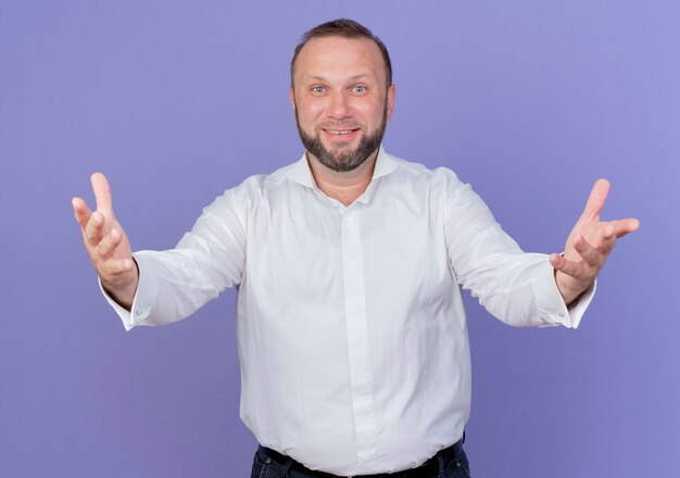 Bärtiger Mann, der weißes Hemd trägt und lächelnde freundliche weit öffnende Hände begrüßt, die über blauer Wand stehend begrüßen