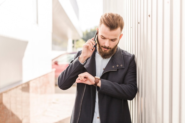 Bärtiger Mann, der Zeit auf Armbanduhr bei der Unterhaltung auf Mobiltelefon betrachtet