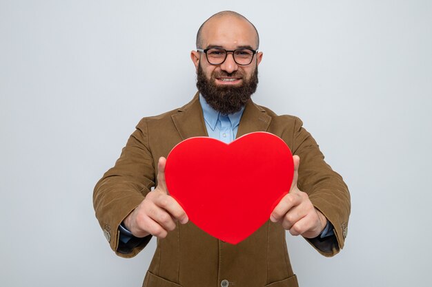 Bärtiger Mann im braunen Anzug mit Brille, der ein Herz aus Pappe hält und fröhlich, glücklich und positiv lächelt