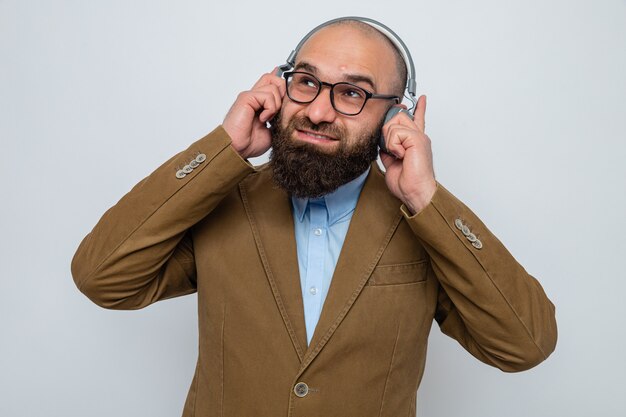 Bärtiger Mann im braunen Anzug mit Brille und Kopfhörern, die lächelnd aufblicken und seine Lieblingsmusik auf weißem Hintergrund genießen