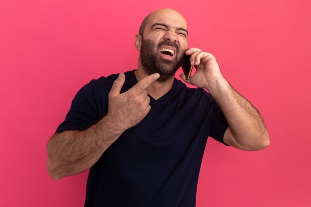 Bärtiger Mann im Marine-T-Shirt, das auf Handy spricht, das frustriert ist, über rosa Wand stehend