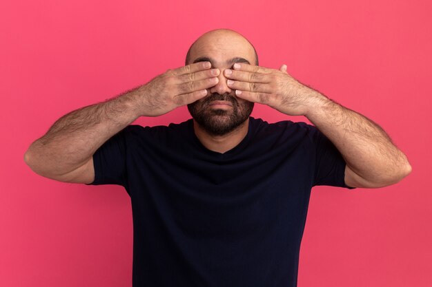 Bärtiger Mann im Marine-T-Shirt, das Augen mit den Händen bedeckt, die über rosa Wand stehen