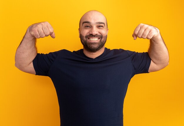 Bärtiger Mann im Marine-T-Shirt, das die Fäuste wie ein glücklicher und aufgeregter lächelnder Gewinner über der orangefarbenen Wand erhebt