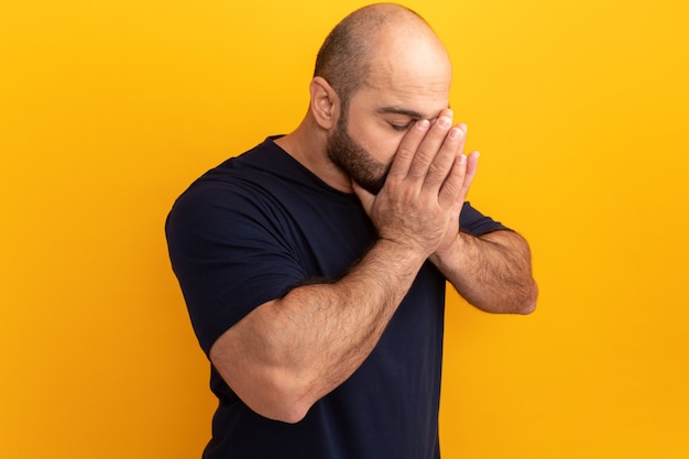 Bärtiger Mann im Marine-T-Shirt, das Hände zusammen auf seinem Gesicht zusammenhält, deprimiert und besorgt, über orange Wand stehend