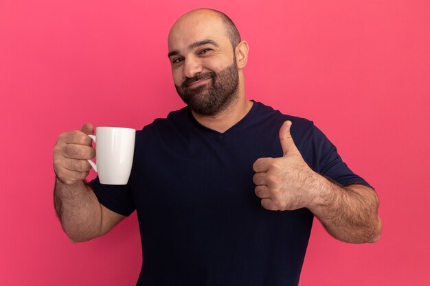 Bärtiger Mann im Marine-T-Shirt, der eine Tasse glücklich und positiv hält, die Daumen oben steht über rosa Wand