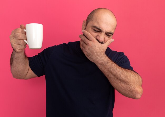 Bärtiger Mann im Marine-T-Shirt, der eine Tasse hält, die müden bedeckenden Mund mit der Hand über rosa Wand steht