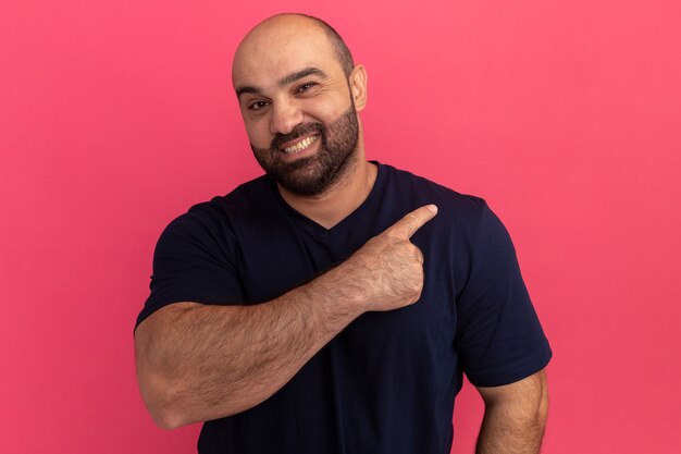 Bärtiger Mann im Marine-T-Shirt, der fröhlich zeigt und zurück zeigt, der über rosa Wand steht