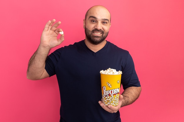 Bärtiger Mann im Marine-T-Shirt-Eimer mit Popcorn mit Lächeln auf Gesicht, das über rosa Wand steht