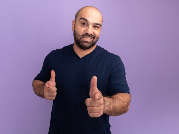 Bärtiger Mann im Marine-T-Shirt glücklich und positiv zeigend mit Zeigefingern, die über lila Wand stehen