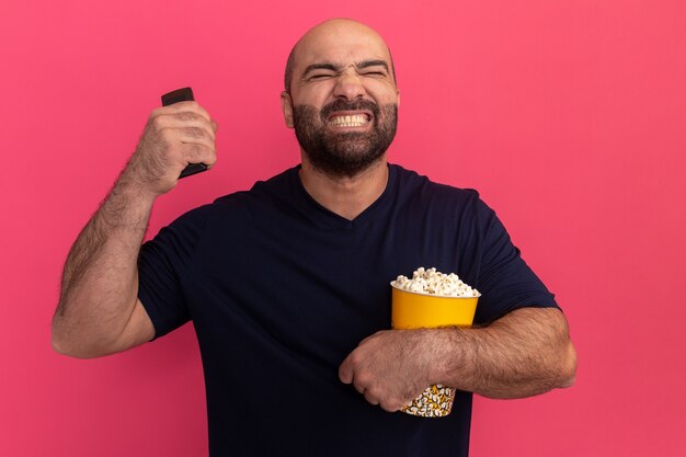 Bärtiger mann im marine-t-shirt hält eimer popcorn und tv-fernbedienung haltend, genervt und gereizt stehend über rosa wand stehend
