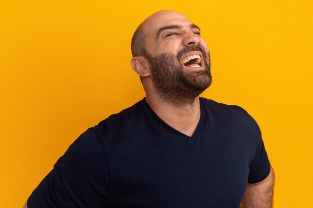 Bärtiger Mann im Marine-T-Shirt lacht glücklich und aufgeregt, über orange Wand stehend