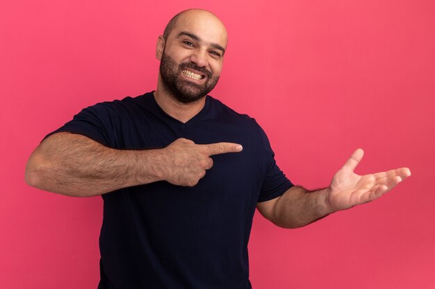Bärtiger Mann im Marine-T-Shirt mit Lächeln auf Gesicht, das Kopienraum mit Arm zeigt, der mit Zeigefinger zur Seite zeigt, die über rosa Wand steht