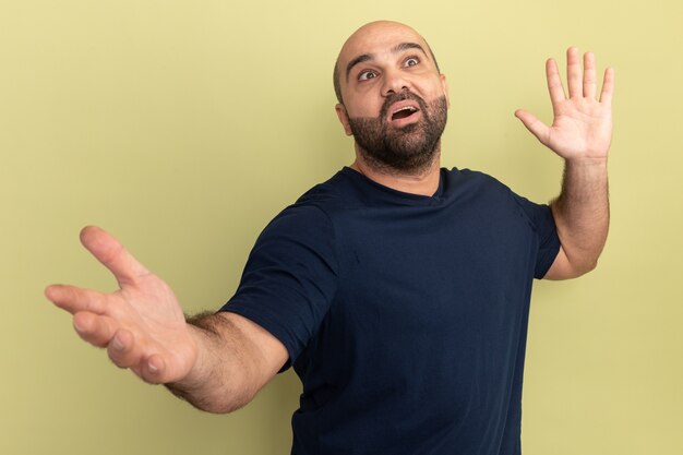 Bärtiger Mann im schwarzen T-Shirt, der lächelnd verwirrte, weit öffnende Hände übergrüner Wand nach oben schaut