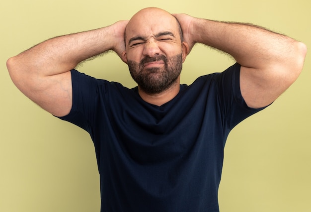 Kostenloses Foto bärtiger mann im schwarzen t-shirt, der verwirrt und genervt mit den händen hinter seinem kopf aussieht, die über grüner wand stehen