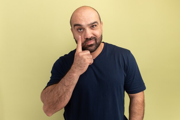 Bärtiger Mann im schwarzen T-Shirt lächelnd mit Zeigefinger auf sein Auge stehend über grüner Wand zeigend