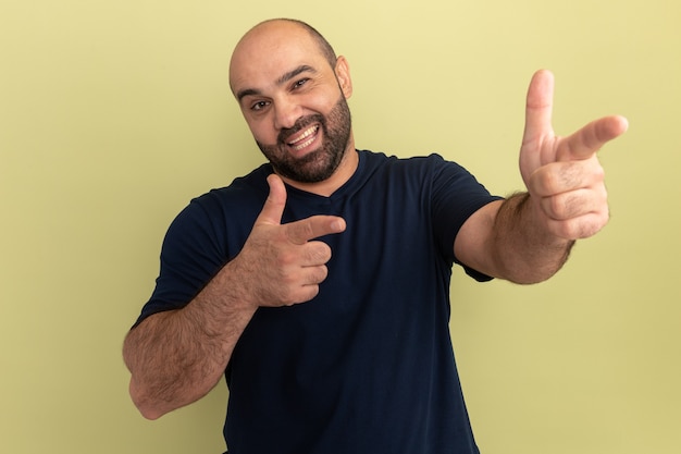 Bärtiger Mann im schwarzen T-Shirt mit dem glücklichen Gesicht, das fröhlich mit Zeigefingern zeigt, die über grüner Wand stehen