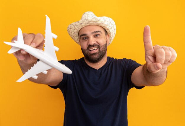 Bärtiger Mann im schwarzen T-Shirt und im Sommerhut, der Spielzeugflugzeug glücklich und fröhlich zeigt Zeigefinger über orange Wand steht