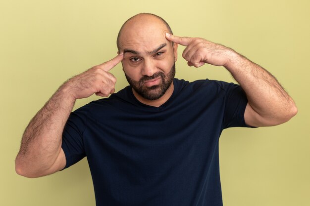 Kostenloses Foto bärtiger mann im schwarzen t-shirt verwirrt mit zeigefinger auf seine schläfen für fehler stehend über grüner wand