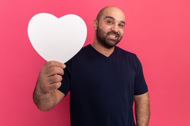 Bärtiger Mann im T-Shirt hält Pappherz lächelnd, der fröhlich über rosa Wand steht