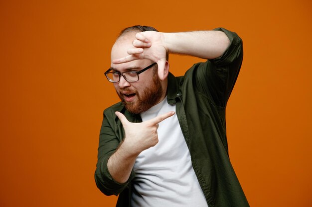 Bärtiger Mann in Freizeitkleidung mit Brille, der einen Rahmen mit Fingern macht, die durch diesen Rahmen in die Kamera schauen, glücklich und fröhlich über orangefarbenem Hintergrund