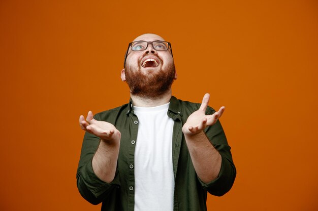 Bärtiger Mann in Freizeitkleidung mit Brille, der glücklich und aufgeregt nach oben blickt und die Arme über orangefarbenem Hintergrund hebt