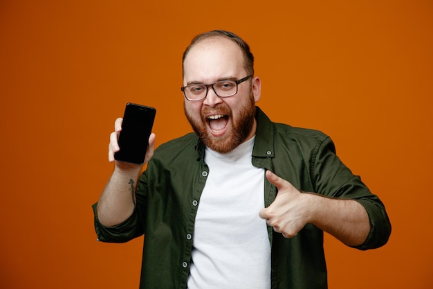 Bärtiger Mann in lässiger Kleidung mit Brille, der das Smartphone zeigt, das glücklich und aufgeregt in die Kamera schaut und den Daumen nach oben zeigt, der über orangefarbenem Hintergrund steht
