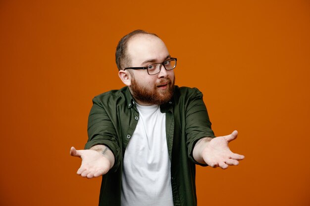Bärtiger Mann in lässiger Kleidung mit Brille, der in die Kamera blickt und Händchen vor ihm hält, als würde er etwas anbieten, das über orangefarbenem Hintergrund steht
