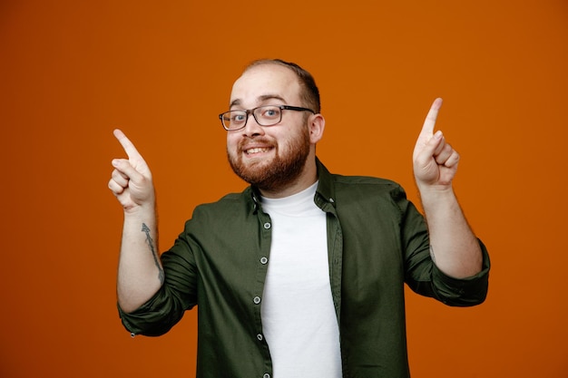 Bärtiger Mann in legerer Kleidung mit Brille, der in die Kamera blickt und fröhlich mit erhobenen Zeigefingern auf orangefarbenem Hintergrund zeigt
