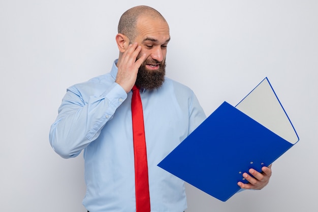 Bärtiger Mann in roter Krawatte und blauem Hemd, der einen Büroordner hält und ihn erstaunt und überrascht auf weißem Hintergrund betrachtet