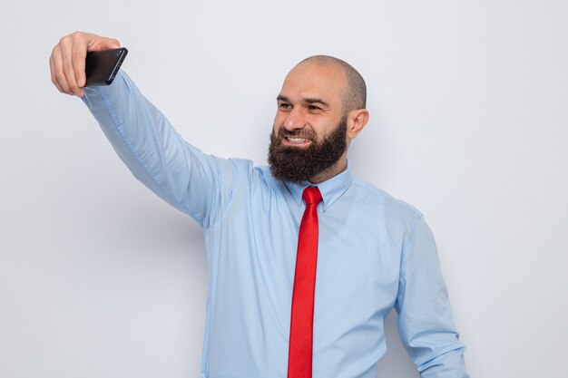 Bärtiger Mann in roter Krawatte und blauem Hemd macht Selfie mit Smartphone und lächelt fröhlich glücklich und positiv