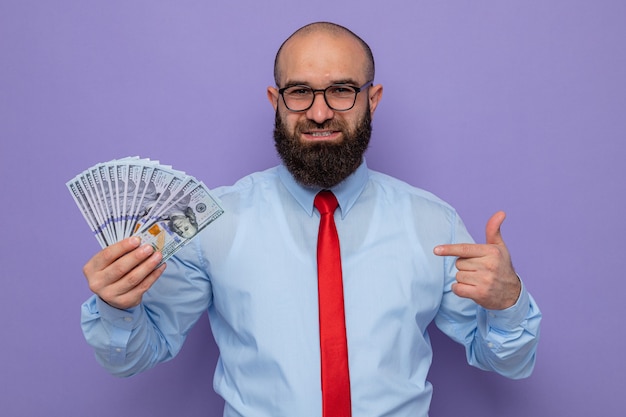 Bärtiger Mann in roter Krawatte und blauem Hemd mit Brille, der Bargeld hält und mit dem Zeigefinger auf Geld zeigt, das auf die Kamera blickt, die fröhlich über lila Hintergrund lächelt