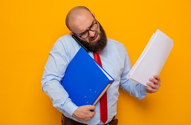 Bärtiger Mann in roter Krawatte und blauem Hemd mit Brille, der Büroordner und Dokumente hält, beschäftigt und gestresst mit dem Handy telefonieren mobile