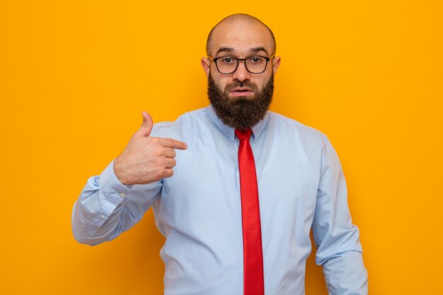 Bärtiger Mann in roter Krawatte und blauem Hemd mit Brille, der die Kamera anschaut, überrascht, mit dem Zeigefinger auf sich selbst zu zeigen, der über orangefarbenem Hintergrund steht