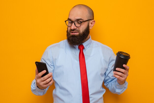 Bärtiger Mann in roter Krawatte und blauem Hemd mit Brille, der Smartphone und Kaffeetasse hält, glücklich und positiv lächelnd, zuversichtlich, über orangefarbenem Hintergrund stehend