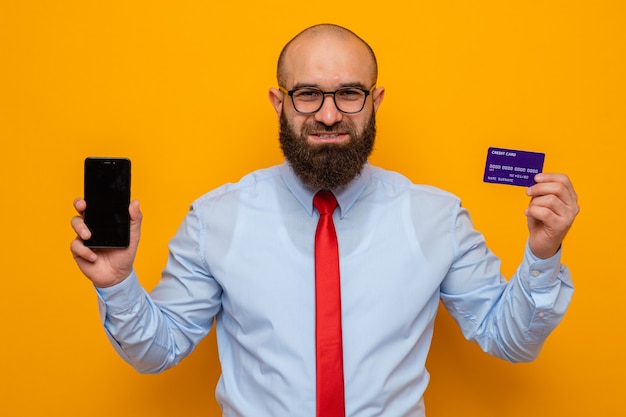 Bärtiger Mann in roter Krawatte und blauem Hemd mit Brille, der Smartphone und Kreditkarte hält und glücklich und positiv lächelt selbstbewusst aussieht