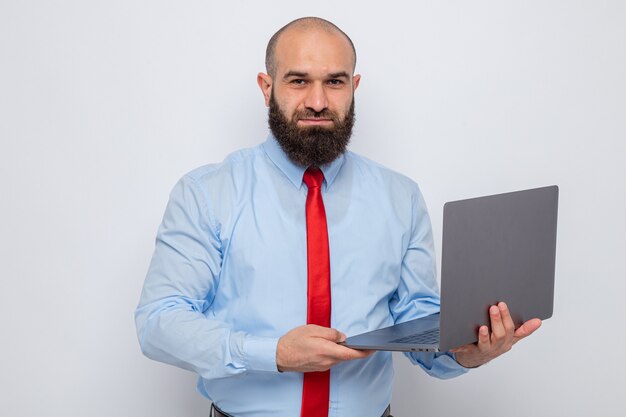 Bärtiger Mann in roter Krawatte und blauem Hemd mit Laptop und Blick in die Kamera lächelt selbstbewusst glücklich und positiv auf weißem Hintergrund