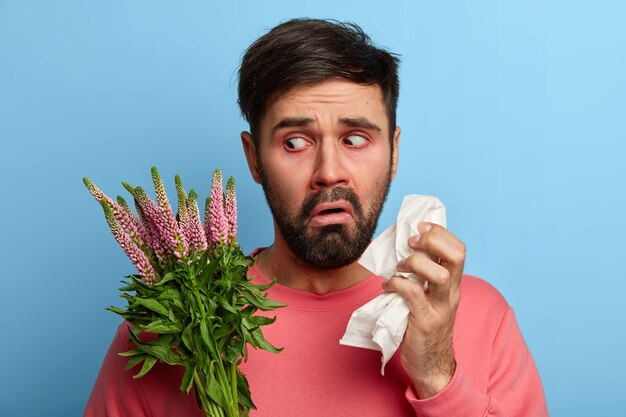 bärtiger Mann leidet an allergischer Rhinitis, hält Serviette und schaut unglücklich auf Allergene, fühlt sich krank, hat laufende Nase und ständiges Niesen, braucht wirksame Medikamente, um Krankheiten zu heilen