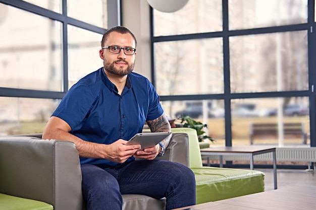 Bärtiger Mann mit Brille mit Tätowierung auf dem Arm sitzt auf einem Stuhl und benutzt einen Tablet-PC.