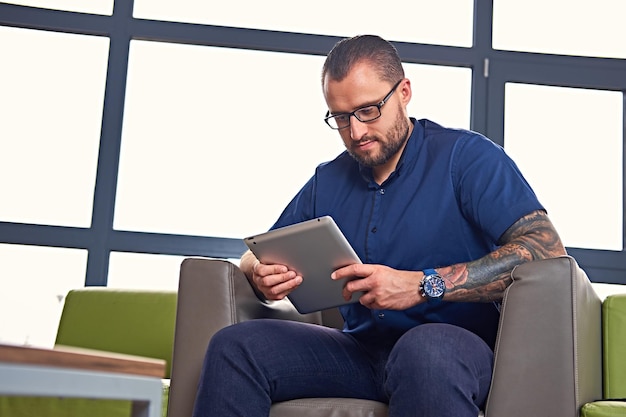 Bärtiger Mann mit Brille mit Tätowierung auf dem Arm sitzt auf einem Stuhl und benutzt einen Tablet-PC.