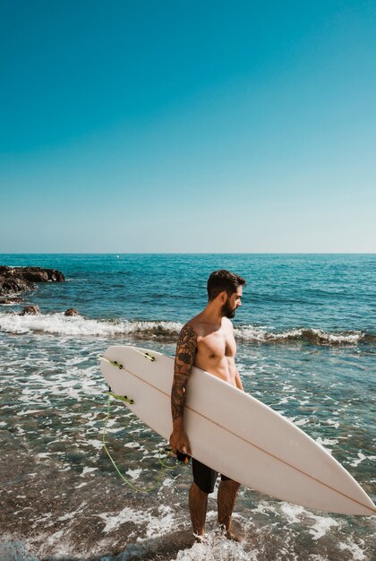 Bärtiger Mann mit dem Surfbrett, das wellenartig bewegendes Meer betrachtet