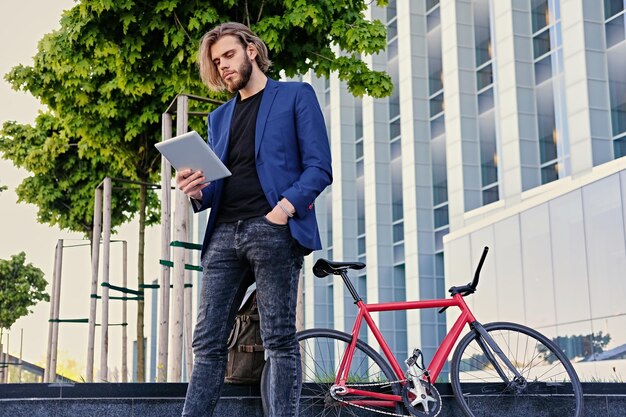 bärtiger Mann mit langen blonden Haaren hält Tablet-PC mit rotem Single-Speed-Fahrrad in einem Park im Hintergrund.