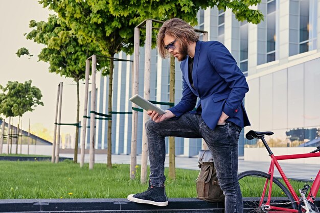 bärtiger Mann mit langen blonden Haaren hält Tablet-PC mit rotem Single-Speed-Fahrrad in einem Park im Hintergrund.