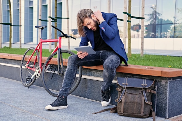 Kostenloses Foto bärtiger mann mit langen blonden haaren hält tablet-pc mit rotem single-speed-fahrrad in einem park im hintergrund.